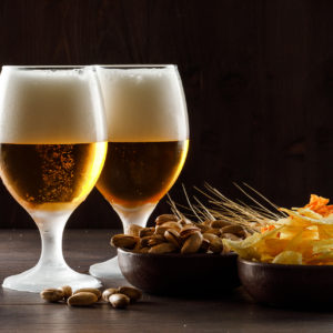 Foamed beer with pistachio, wheat ears, chips in goblet glasses on wooden background, side view.